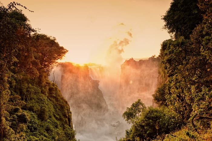 Sunset over Victoria Falls framed by lush green foliage and rising mist