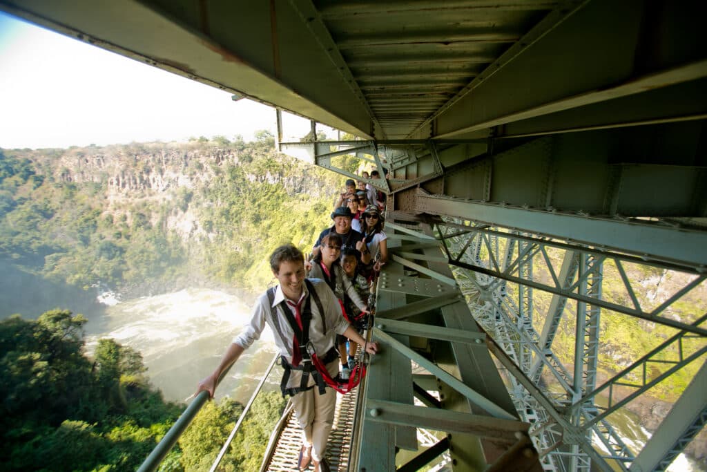 A group of tourists in safety harnesses walk along a narrow metal walkway under a large bridge, high above a river canyon