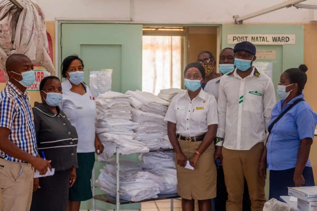 A group of healthcare workers and staff wearing face masks pose in front of stacks of white linens outside a hospital ward