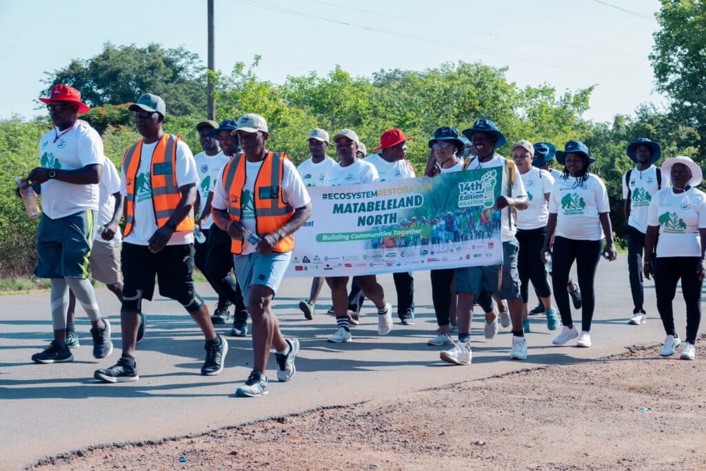A group of people wearing matching white shirts walk down a paved road, carrying a banner that reads "14th Edition Matabeleland North Walkathon