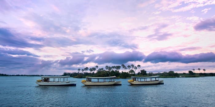 Three covered boats on a calm river under a purple and pink sunset sky