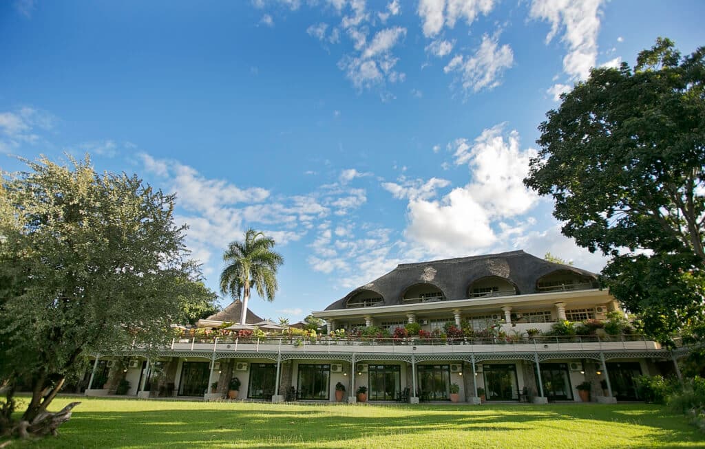 A two-story resort building with a thatched roof stands beside a grassy lawn under a blue sky with scattered clouds