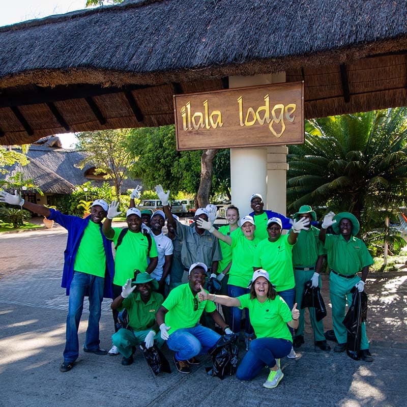 A group of smiling staff and volunteers in green shirts pose with trash bags and gloves in front of the Ilala Lodge entrance