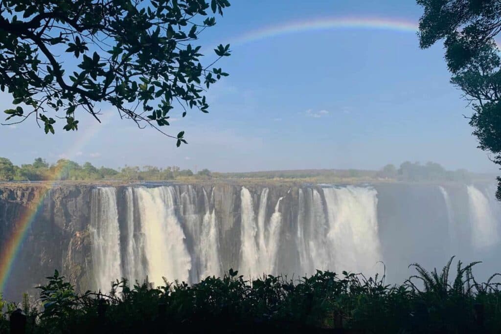 A wide waterfall cascading over a cliff, framed by lush green trees and a faint rainbow in the sky above