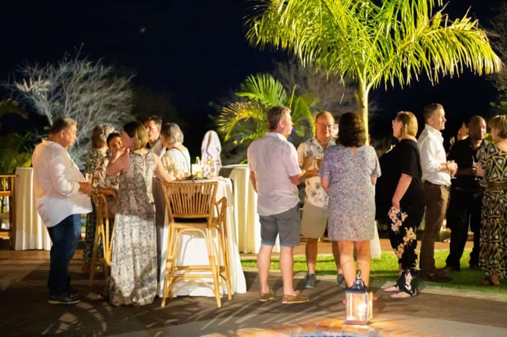 People socializing outdoors at night around cocktail tables lit by lanterns and palm trees