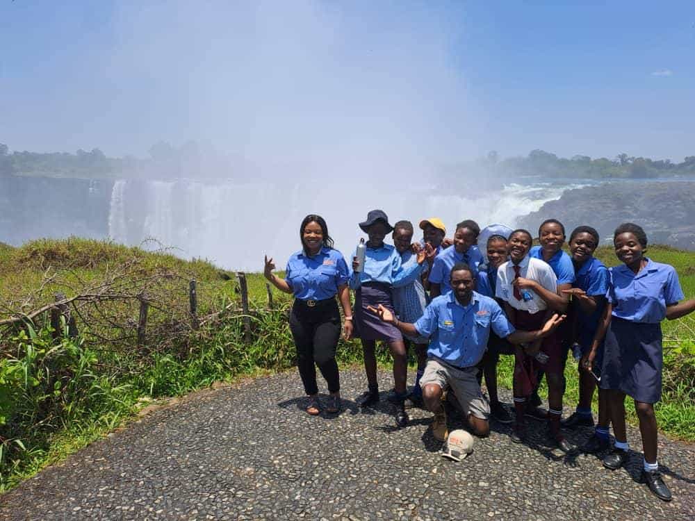 A group of students and two adults pose happily on a stone path with a misty waterfall in the background