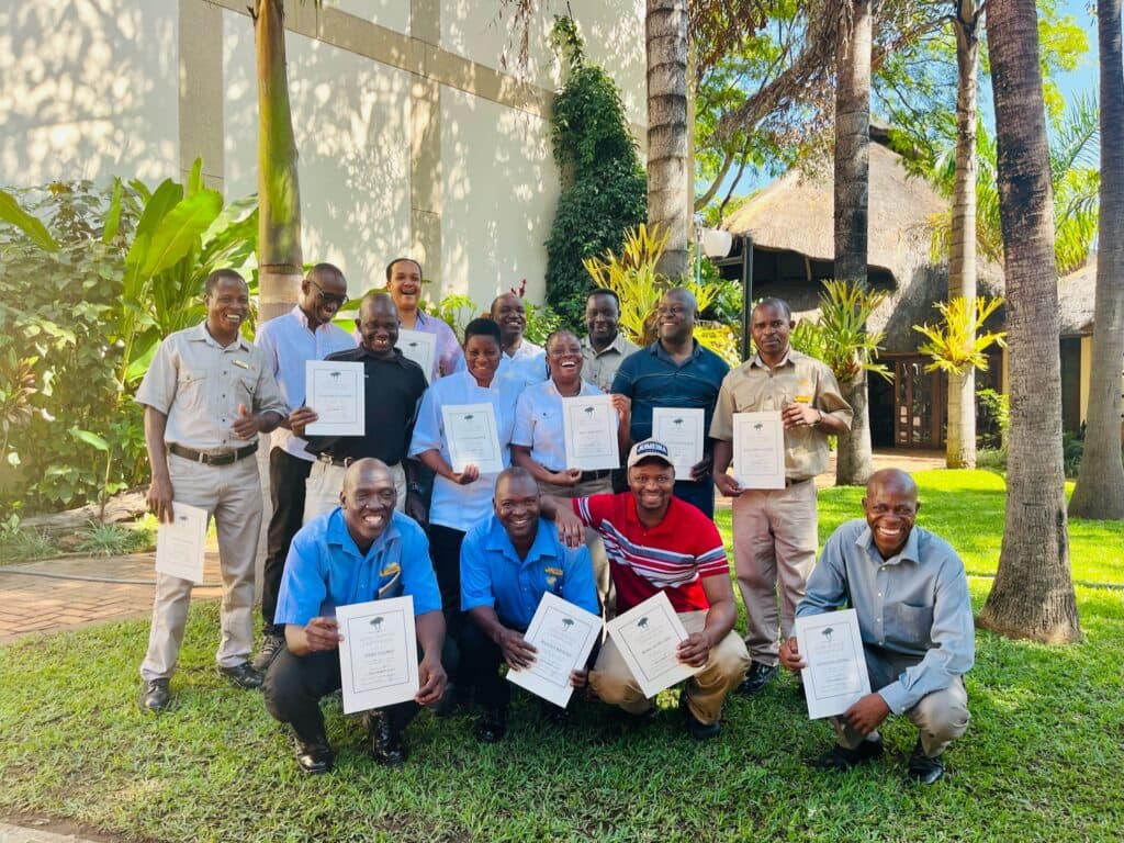 A group of sixteen smiling staff members outdoors on a lawn, holding up certificates