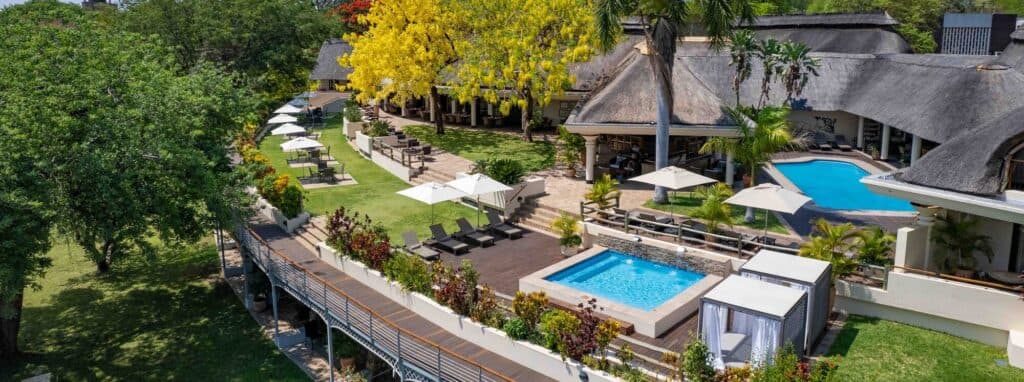 An aerial view of a resort with thatched-roof buildings, swimming pools, lounge chairs, umbrellas, and lush green grounds
