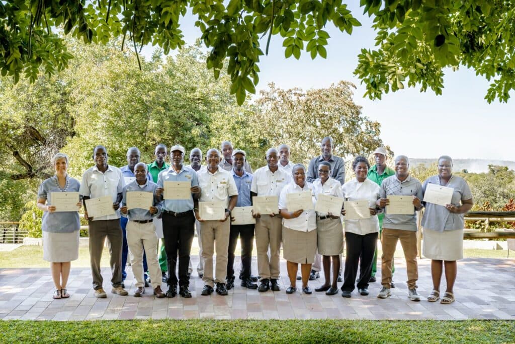 Group of nineteen staff members standing outdoors on a paved patio holding their certificates, framed by trees above