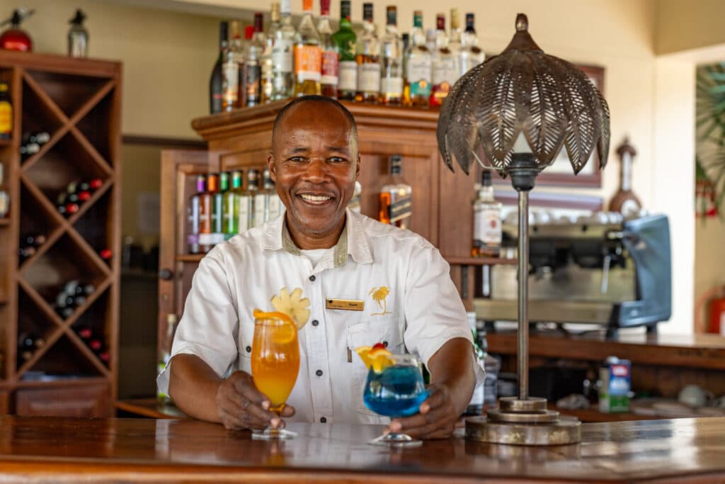 A bartender smiling behind a bar, holding up two colorful cocktails
