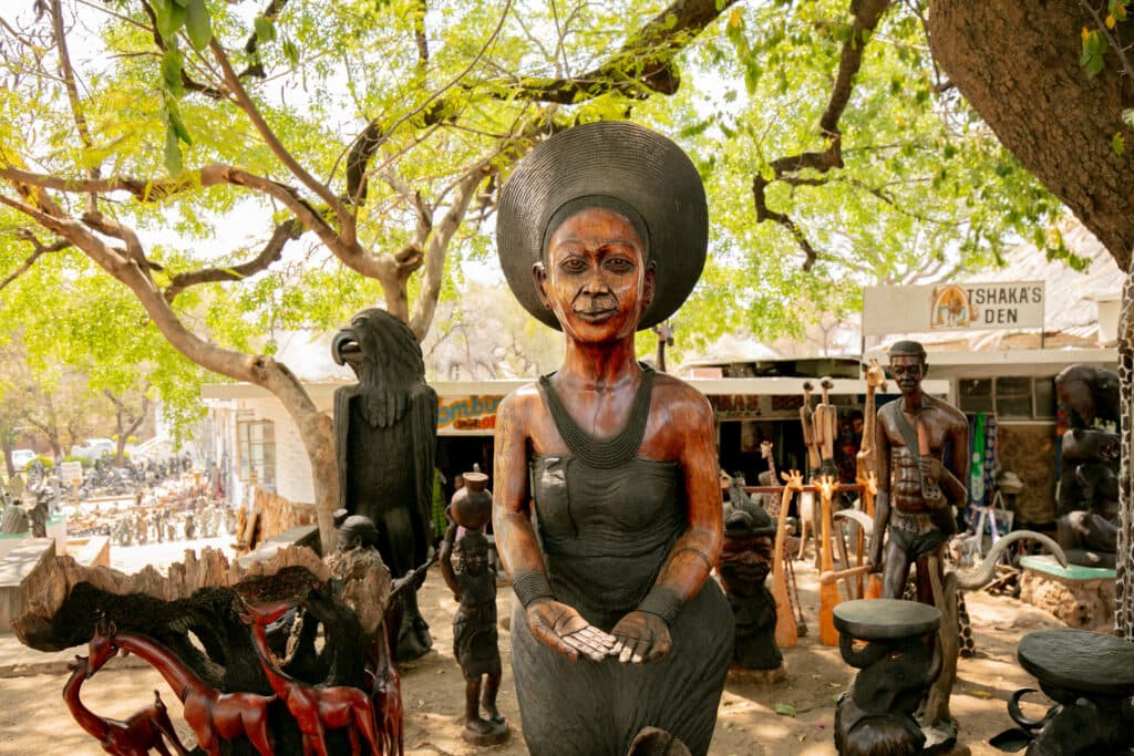 Wooden carved statues on display outdoors, featuring a life-sized woman wearing a wide hat and cupping her hands