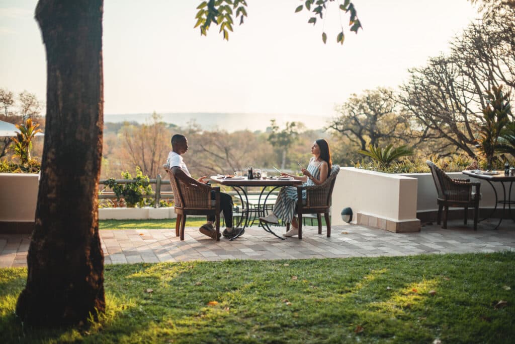 A man and woman enjoy an outdoor meal on a sunny patio overlooking a landscape