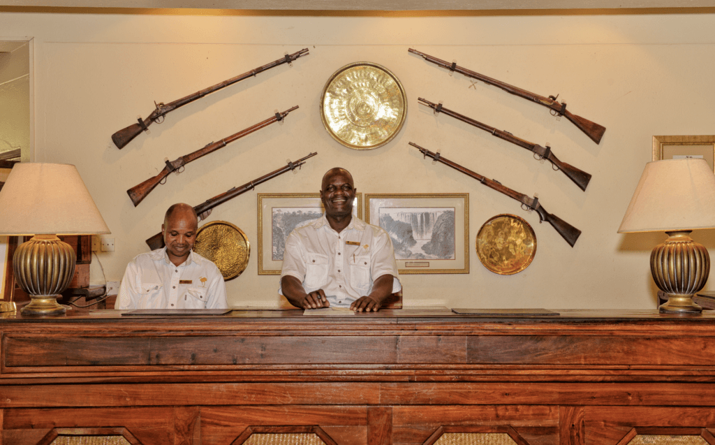 Two smiling male staff members stand behind a wooden reception desk beneath a wall decorated with historical rifles and framed pictures